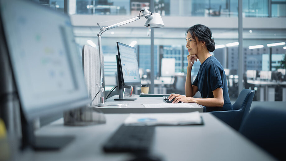 Frau sitzt in einem großen offenen Büro vor einem Laptop.