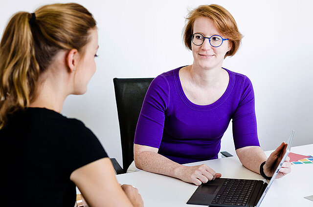 Zwei Frauen sitzen an einem Bürotisch mit aufgeklapptem Laptop und unterhalten sich.