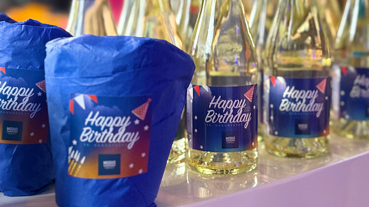Ein Geburtstagskuchen im Glas steht neben mehreren Sektfläschen, die kleine bunte Sticker mit der Aufschrift " Happy Birthday MODUS Consult" tragen. 