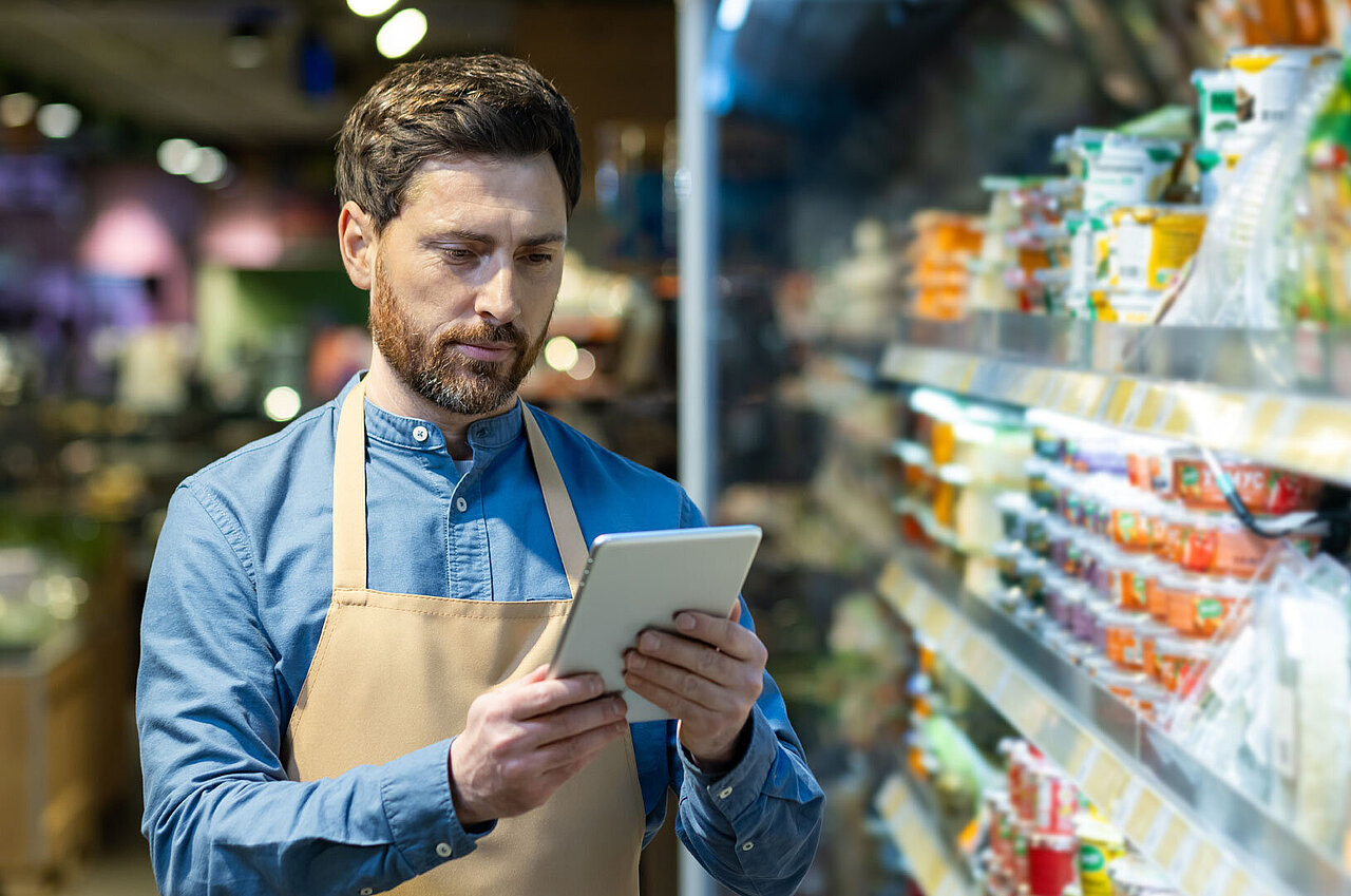 Sympathischer Mann mit Tablet in der Hand, der vor einem Lebensmittelregal in einem Supermarkt steht. 