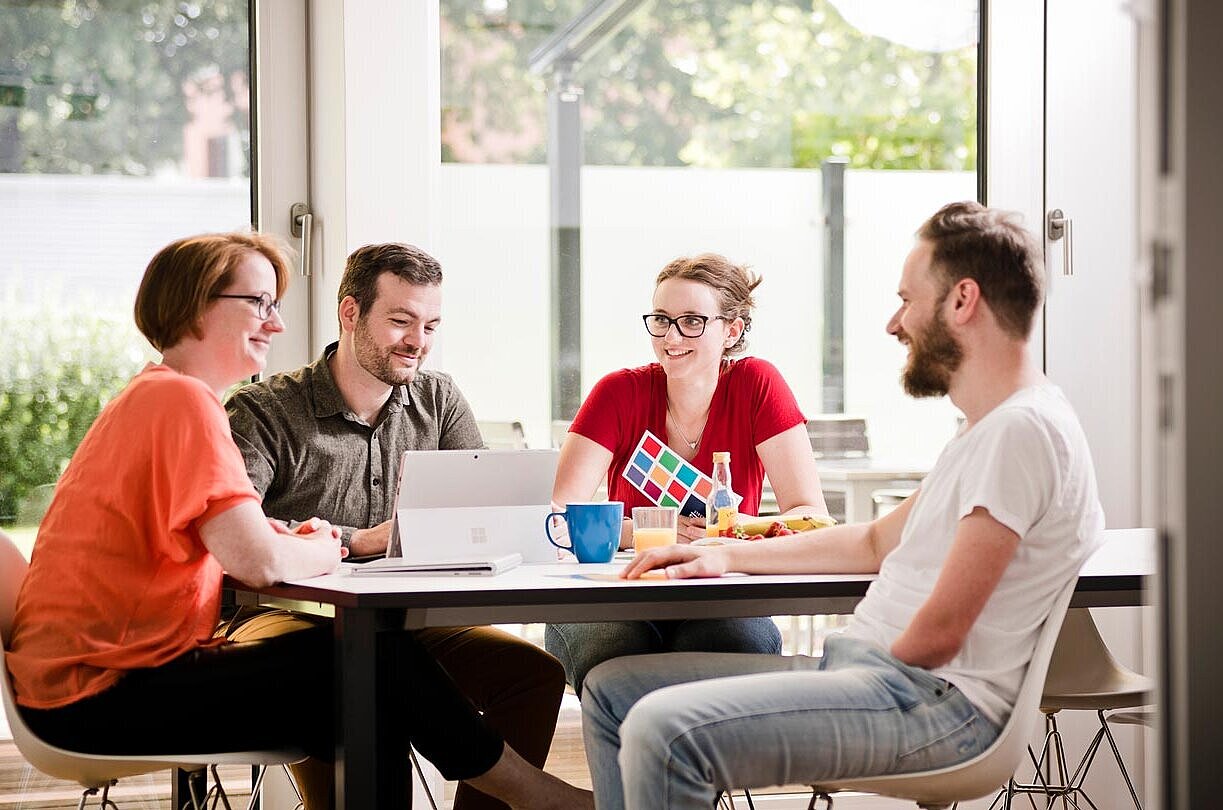 Vier Kollegen bei MODUS Consult, die locker an einem Tisch gemeinsam sitzen und sich unterhalten.